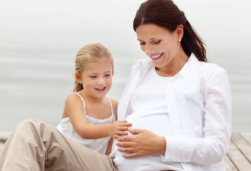 Kobieta razem z córeczką siedzą na pomoście i głaszczą brzuch ciążowy mamy