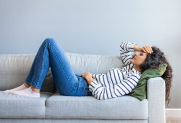 Zmęczona kobieta leży na kanapie i trzyma się za głowę.